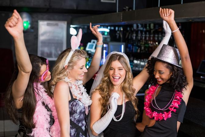 Four friends at a bar celebrating a bachelorette party, laughing and dancing in veils, bunny ears, feather boas and a glitter hat.