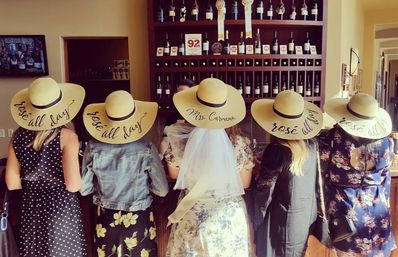 Five friends in floppy straw hats that say "rosé all day" standing at a winery tasting room bar with shelves of wine bottles — center wearing a bridal veil for a playful bachelorette rosé tasting.