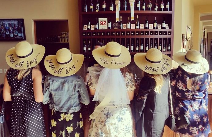 Five friends in floppy straw hats that say "rosé all day" standing at a winery tasting room bar with shelves of wine bottles — center wearing a bridal veil for a playful bachelorette rosé tasting.