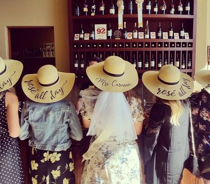 Five friends in floppy straw hats that say "rosé all day" standing at a winery tasting room bar with shelves of wine bottles — center wearing a bridal veil for a playful bachelorette rosé tasting.