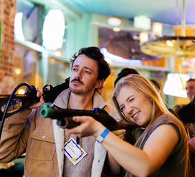 Two friends at a neon-lit indoor arcade bar playing a rifle-shooter game — woman squinting to aim, man posing casually in a lively night-out scene