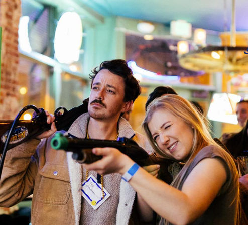 Two friends at a neon-lit indoor arcade bar playing a rifle-shooter game — woman squinting to aim, man posing casually in a lively night-out scene