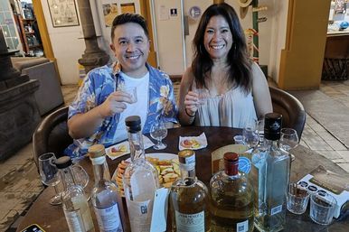 Two smiling people enjoying a tequila tasting indoors, holding small tasting glasses at a table filled with multiple tequila bottles, shot glasses, and snack plates.