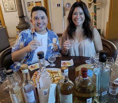Two smiling people enjoying a tequila tasting indoors, holding small tasting glasses at a table filled with multiple tequila bottles, shot glasses, and snack plates.