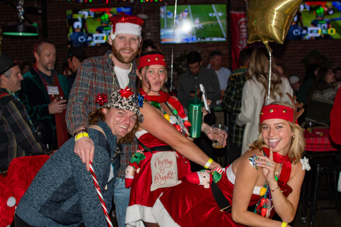 Four friends in playful Christmas costumes and accessories posing and laughing at a crowded sports bar holiday party with football on TV