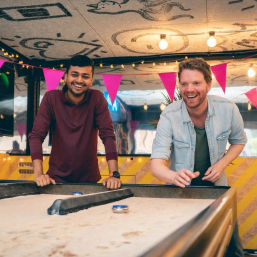 Two men laughing and playing shuffleboard at a festive indoor game area with pink bunting, string lights, and decorative ceiling art.