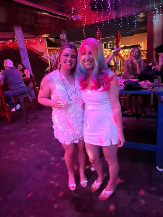 Two friends in white dresses smiling and posing at a lively outdoor bar at night under pink and purple string lights; one wears a sash and holds a drink, festive nightlife scene with patrons and tables in the background.