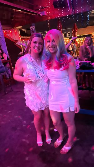 Two friends in white dresses smiling and posing at a lively outdoor bar at night under pink and purple string lights; one wears a sash and holds a drink, festive nightlife scene with patrons and tables in the background.
