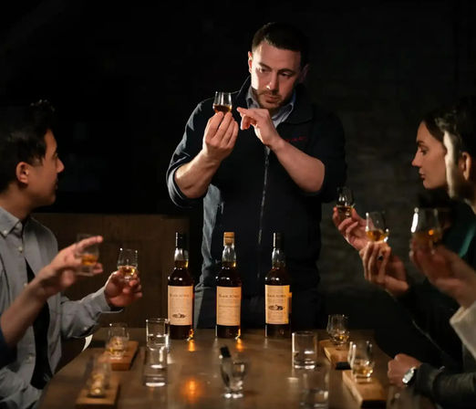 Whiskey tasting: a guide holds a nosing glass up for inspection while four guests sample pours around a wooden table with three bottles in a dimly lit tasting room.