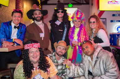 Group of eight adults in playful costumes—cowboy, hippie, rocker with top hat, glitter jacket and rainbow feather outfit—smiling and posing together at an indoor bar during a lively costume party