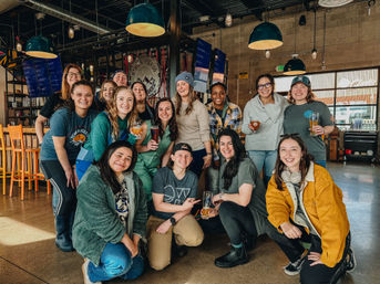 Cheerful group of friends posing with beers in a sunlit craft brewery taproom with industrial pendant lights, wooden bar, and casual downtown vibe.