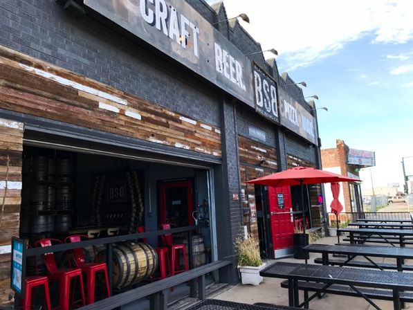 Urban craft brewery exterior with open garage entrance, reclaimed wood facade, stacked beer barrels, red metal stools, red door and umbrella, and outdoor picnic-table seating on a sunny day.