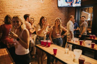 Lively group of friends playing beer pong at a brick‑walled bar, wooden tables stacked with red plastic cups and pitchers in a casual indoor nightlife scene.