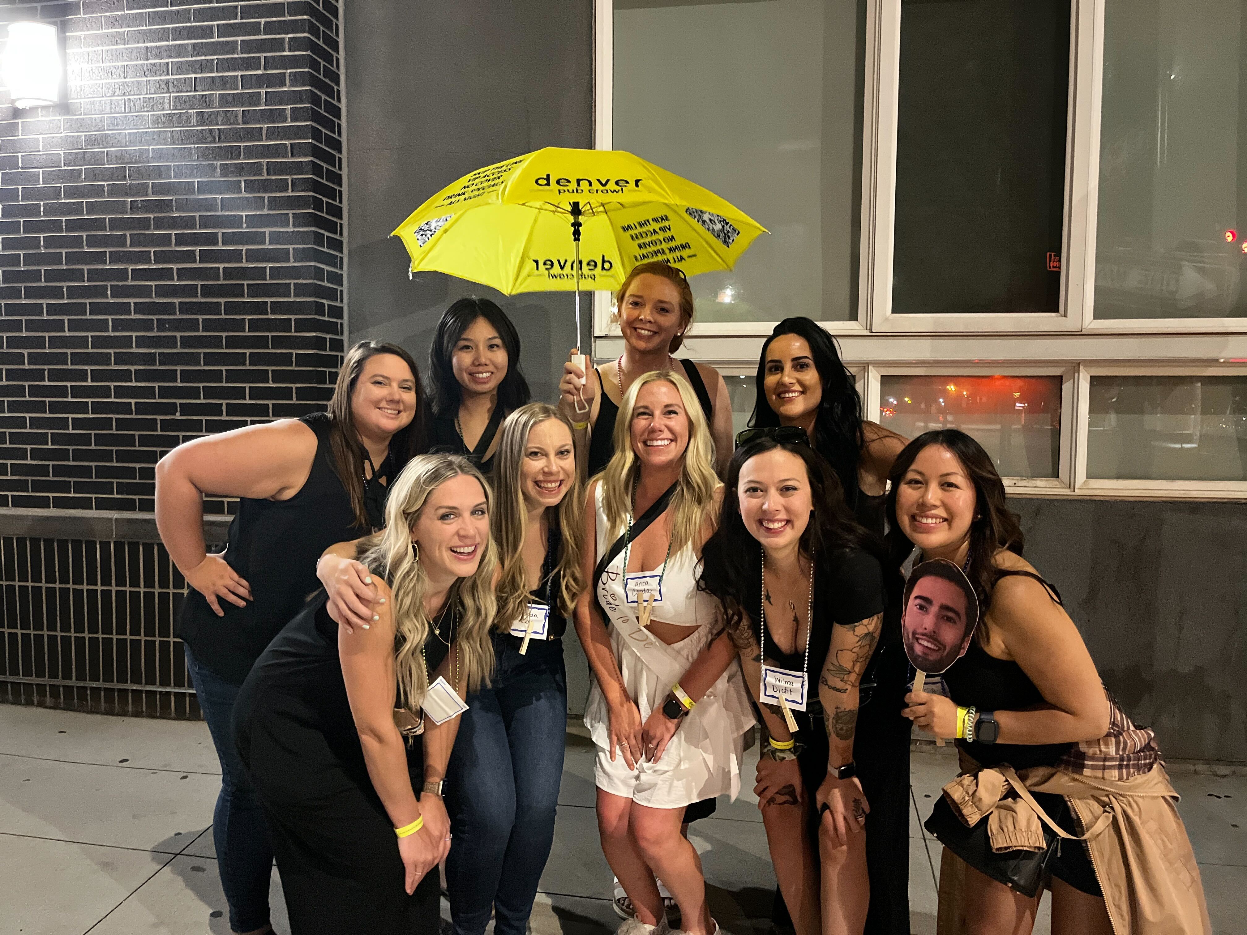 Cheerful group of women posing at night outside a brick building in Denver, one holding a bright yellow umbrella and a woman wearing a bachelorette sash