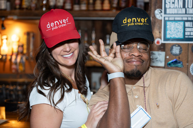 Two smiling adults in event caps playfully posing by beer taps at a lively Denver bar
