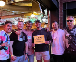 Six friends posing and smiling in a downtown Denver bar during a pub crawl, wearing name tags and beads; one holds a yellow event sign under warm ceiling lights.