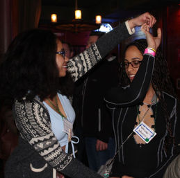 Two friends dancing at a dimly lit bar or lounge, smiling and holding hands; one wears a patterned cardigan and blue top, the other a black laced-front top with name tags and beaded necklaces for a lively nightlife scene.