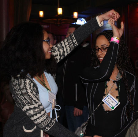 Two friends dancing at a dimly lit bar or lounge, smiling and holding hands; one wears a patterned cardigan and blue top, the other a black laced-front top with name tags and beaded necklaces for a lively nightlife scene.