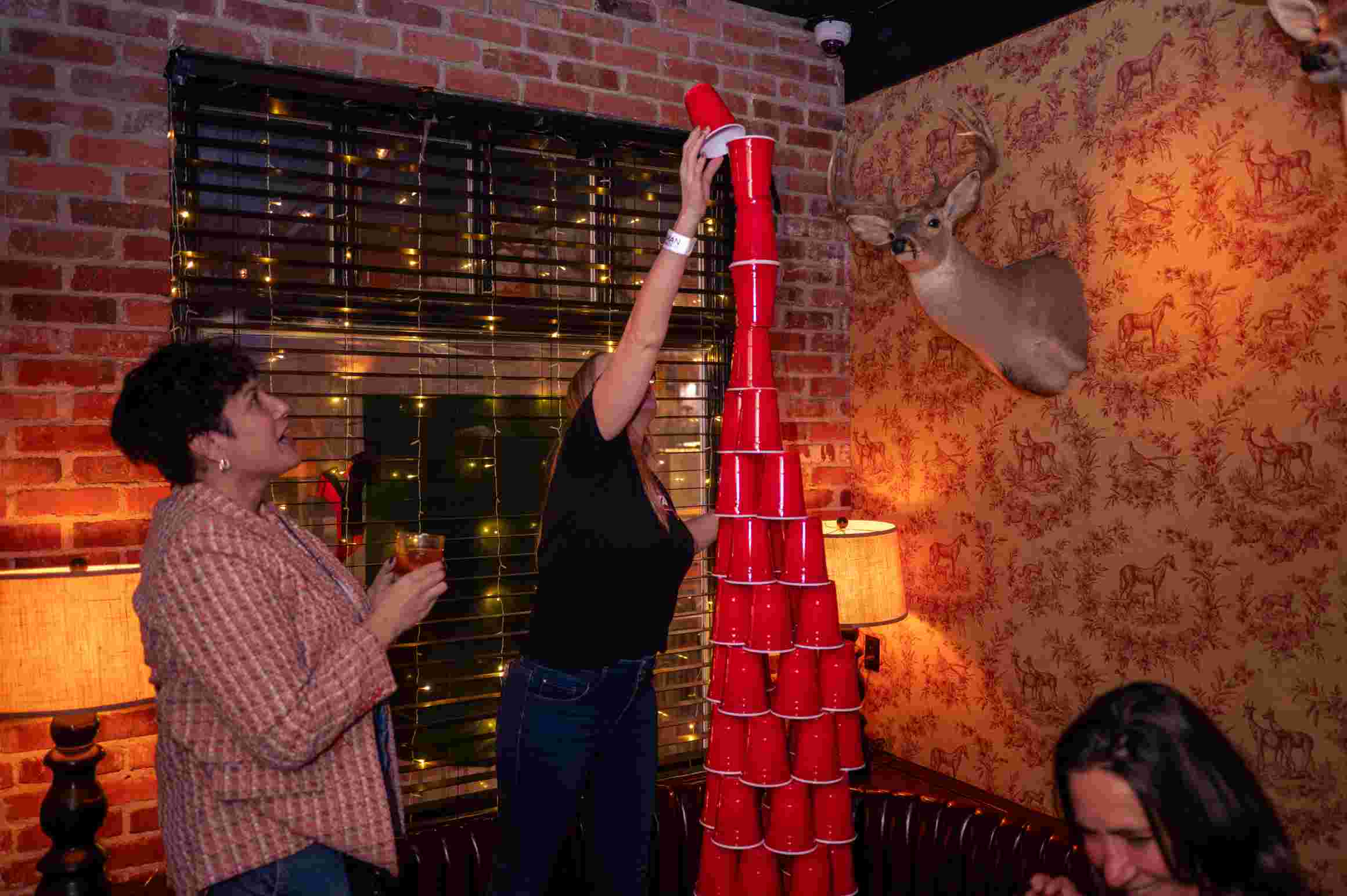 Three people stack a tall tower of red solo cups in a cozy brick‑walled bar with warm lamps, string lights and a mounted deer head on patterned wallpaper.