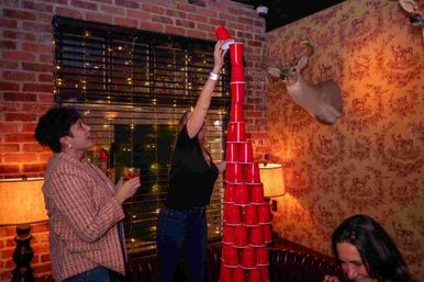 Three people stack a tall tower of red solo cups in a cozy brick‑walled bar with warm lamps, string lights and a mounted deer head on patterned wallpaper.