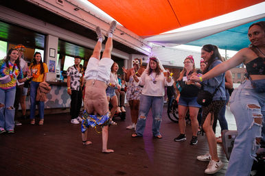 Man doing a handstand on a covered outdoor deck at a lively summer rooftop-bar party, friends cheering and holding drinks under colorful shade sails