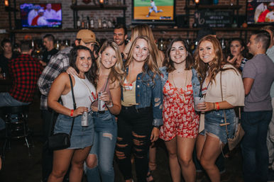 Group of five women smiling and holding drinks in a lively bar with exposed brick walls and TV screens — fun night out, city nightlife scene.