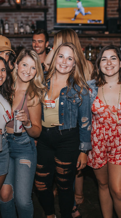 Group of five women smiling and holding drinks in a lively bar with exposed brick walls and TV screens — fun night out, city nightlife scene.