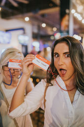 Two women at a lively bar party holding 'Hello my name is' badge necklaces—one making a surprised face and showing a humorous 'Mike Oxsmall' name tag amid warm bokeh lights.
