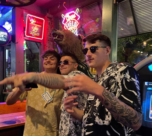 Three men in Hawaiian shirts and sunglasses pose and point under neon signs beside a mounted bear head inside a neon-lit bar — lively downtown nightlife scene