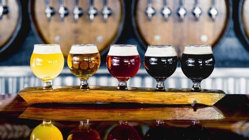 Vibrant craft beer flight of five snifter glasses on a wooden paddle at a brewery bar, with oak barrels and taps blurred in the background