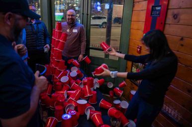 People at a lively indoor bar knocking over a tall tower of red plastic cups as the cups cascade across a table near wood-paneled walls and a glass door.