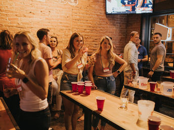 Group of people laughing and playing beer-pong with red plastic cups on wooden tables inside a lively brick‑walled bar with TVs — casual urban nightlife scene.