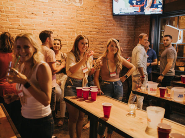 Group of people laughing and playing beer-pong with red plastic cups on wooden tables inside a lively brick‑walled bar with TVs — casual urban nightlife scene.