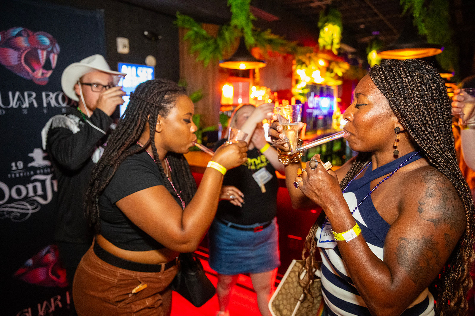 Friends sipping cocktails through a shared long drinking tube in a neon-lit bar, braided hairstyles, tattoos, wristbands and lively nightlife.