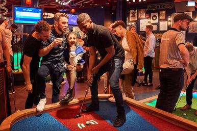 Group of friends at an indoor mini-golf bar, man putting on a colorful carpeted hole while others cheer in a neon-lit arcade and nightlife setting