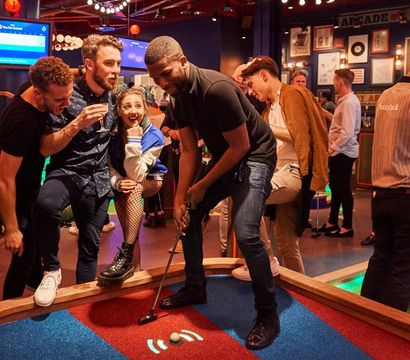 Group of friends at an indoor mini-golf bar, man putting on a colorful carpeted hole while others cheer in a neon-lit arcade and nightlife setting