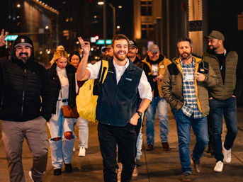 Smiling group of friends walking down a downtown city street at night, lead man waving with a yellow backpack
