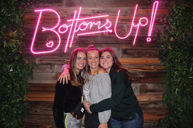 Three smiling women hugging under a pink neon Bottoms Up sign on a rustic wooden backdrop with leafy green accents, lively party group photo