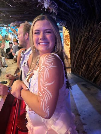 Smiling young woman at a tiki-style bar wearing beads and a white sash, showcasing decorative white floral body paint on her upper arm in a lively nightlife scene.