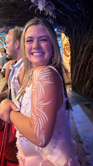 Smiling young woman at a tiki-style bar wearing beads and a white sash, showcasing decorative white floral body paint on her upper arm in a lively nightlife scene.