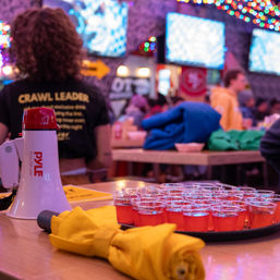 Busy pub crawl scene: tray of red shot cups beside a white megaphone and rolled yellow jacket on a table, with a person in a 'CRAWL LEADER' shirt, colorful string lights and TV screens in the lively bar background.