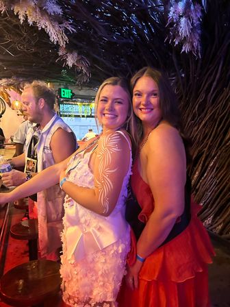 Two smiling women posing in a colorful tropical-themed bar at night — one in a white dress with floral body paint on her arm, the other in a red dress, surrounded by purple lighting and wicker decor.