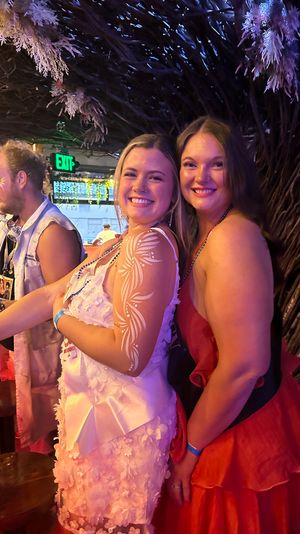Two smiling women posing in a colorful tropical-themed bar at night — one in a white dress with floral body paint on her arm, the other in a red dress, surrounded by purple lighting and wicker decor.
