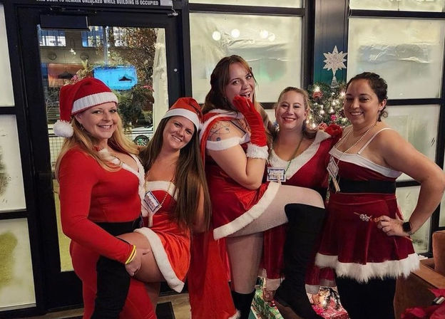 Five women in festive Santa and Mrs. Claus outfits posing cheerfully indoors by a decorated Christmas tree at a holiday party.