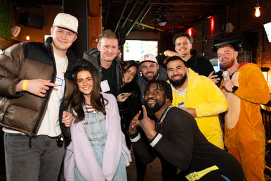Cheerful diverse group of friends posing together inside an urban bar or pub at night, wearing winter jackets and colorful onesies, smiling and holding drinks against a warm brick interior with hanging lights — lively nightlife scene.