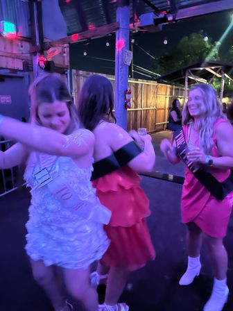 Three women dancing and laughing under purple party lights on an outdoor bar patio at night, wearing colorful dresses and sashes and holding drinks.