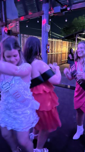 Three women dancing and laughing under purple party lights on an outdoor bar patio at night, wearing colorful dresses and sashes and holding drinks.
