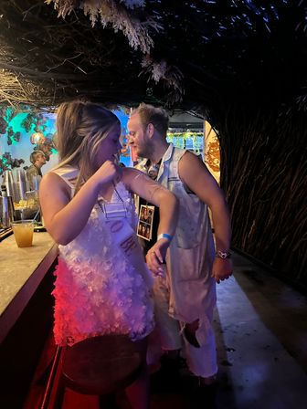 Bachelorette party scene in a dim tropical-themed bar: woman in white floral dress and sash laughing with a man in ripped overalls beneath a woven-branch arch, colorful blue and purple lighting and a cocktail on the bar.