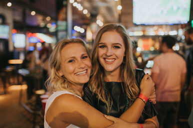 Two smiling young women hugging in a lively sports bar, warm string lights and TV screens glowing in the blurred background — fun downtown nightlife scene.