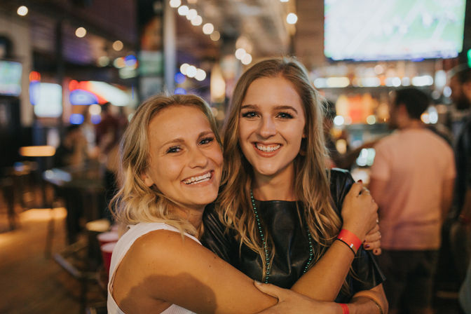 Two smiling young women hugging in a lively sports bar, warm string lights and TV screens glowing in the blurred background — fun downtown nightlife scene.